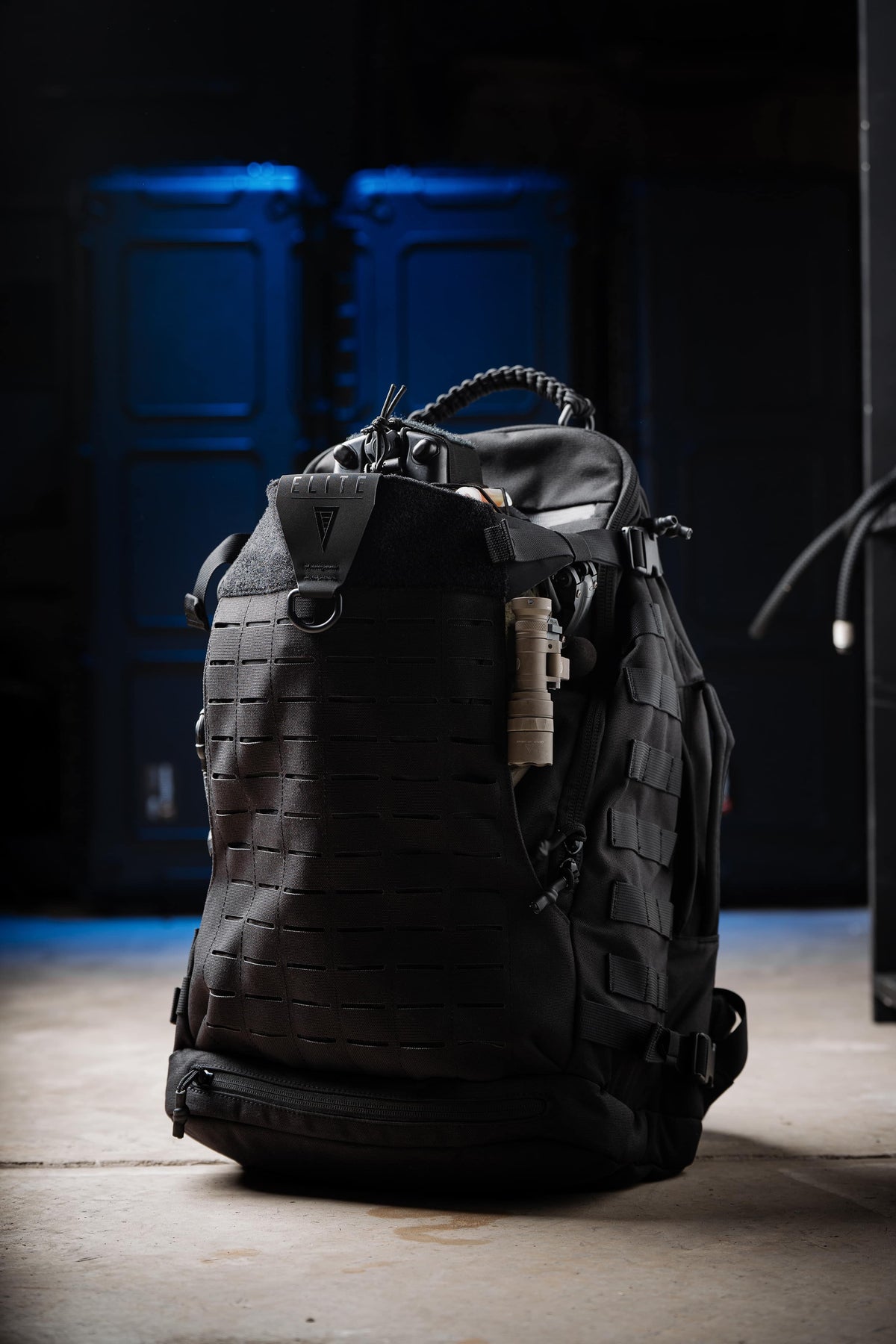 A black tactical backpack with MOLLE webbing and a vertical front pocket, shown on a concrete floor with a dark, moody background featuring blue ambient lighting.