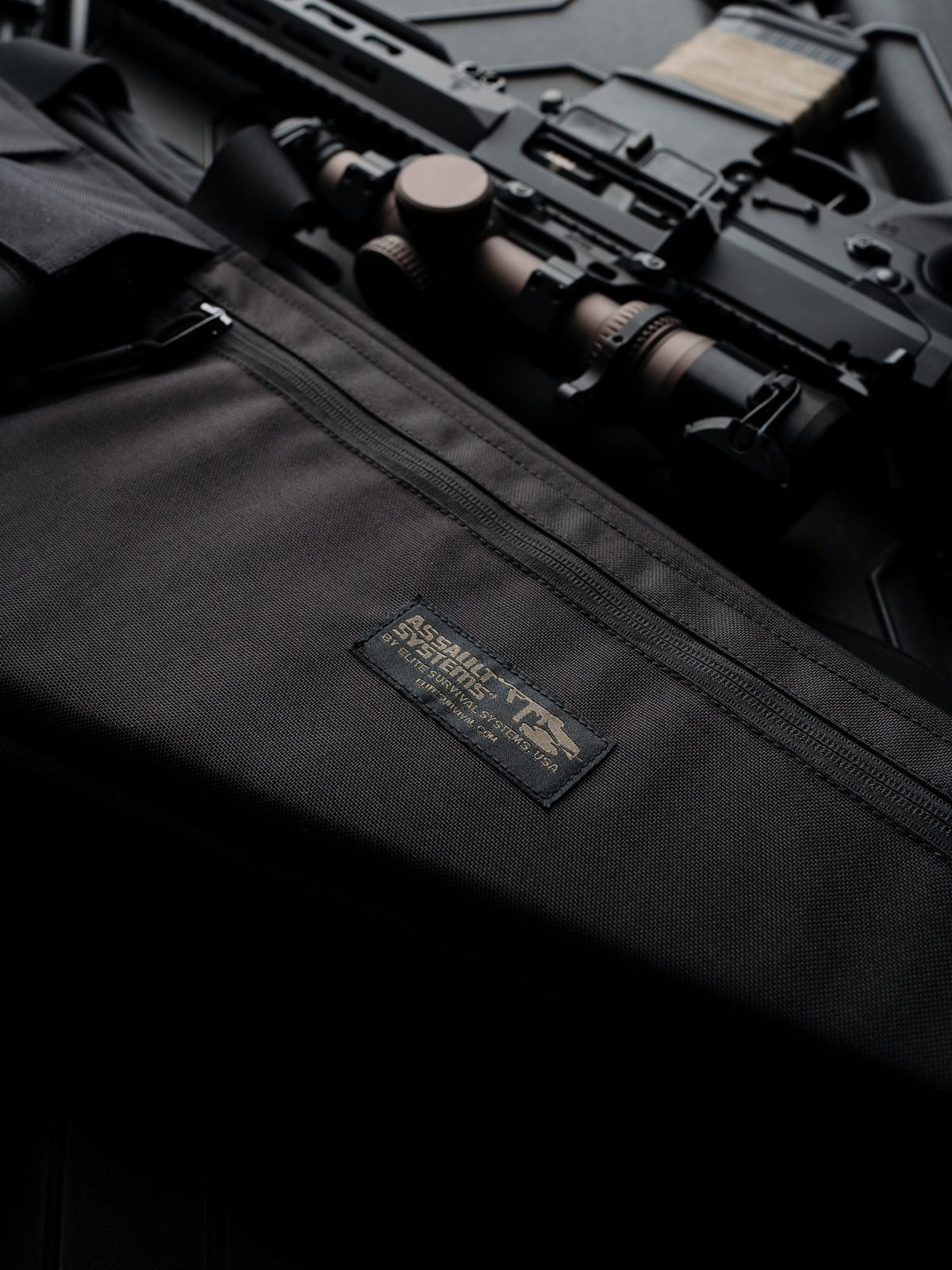 Black Assault Systems rifle case with woven label and zippered pocket, beside a scoped rifle