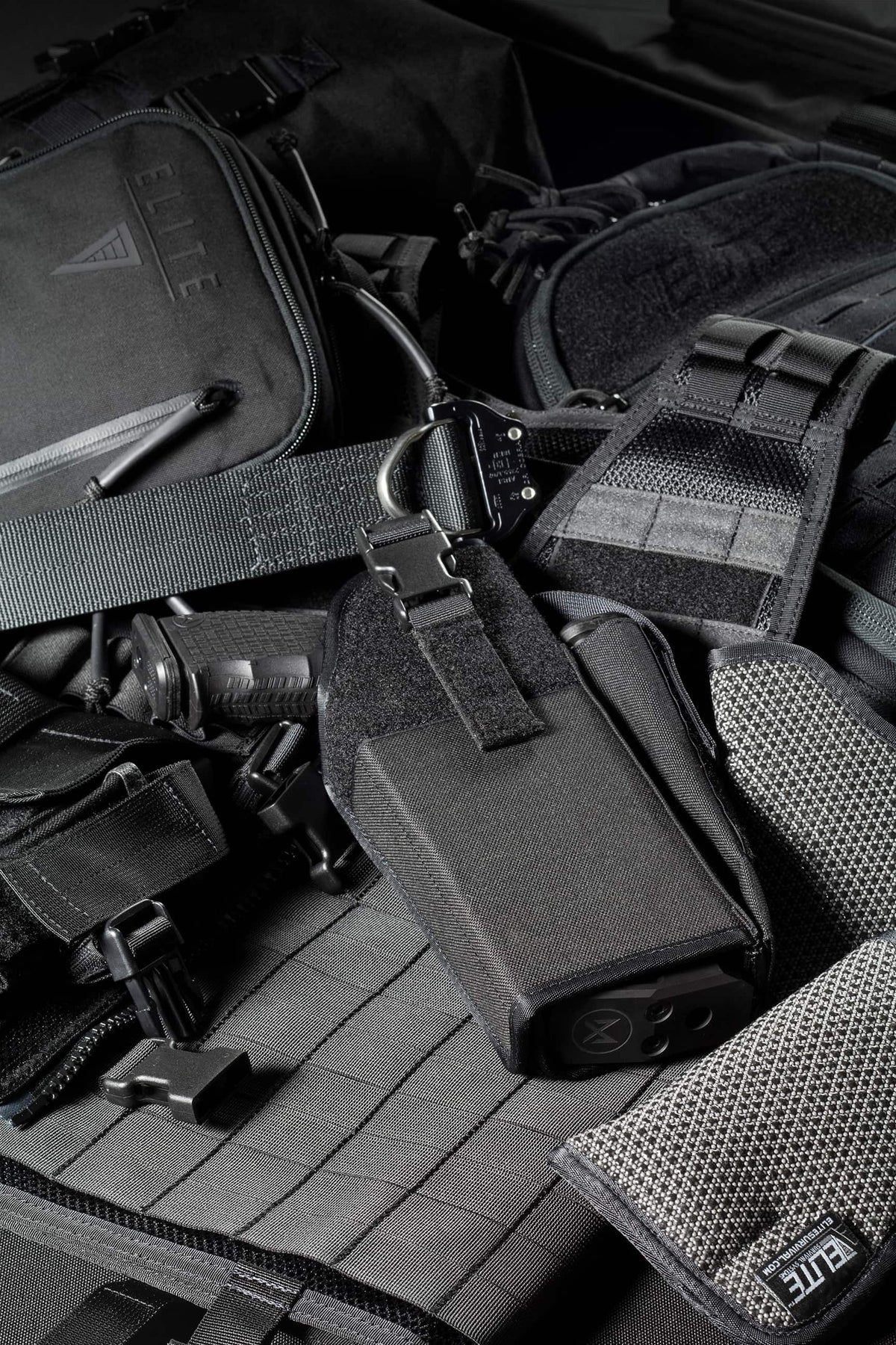 A close-up of tactical gear including a black tactical bag, a black pouch with a metal clip, a gray mesh pocket, and straps laid out on a black tactical mat. The brand name 'ELITE' is visible on some items.
