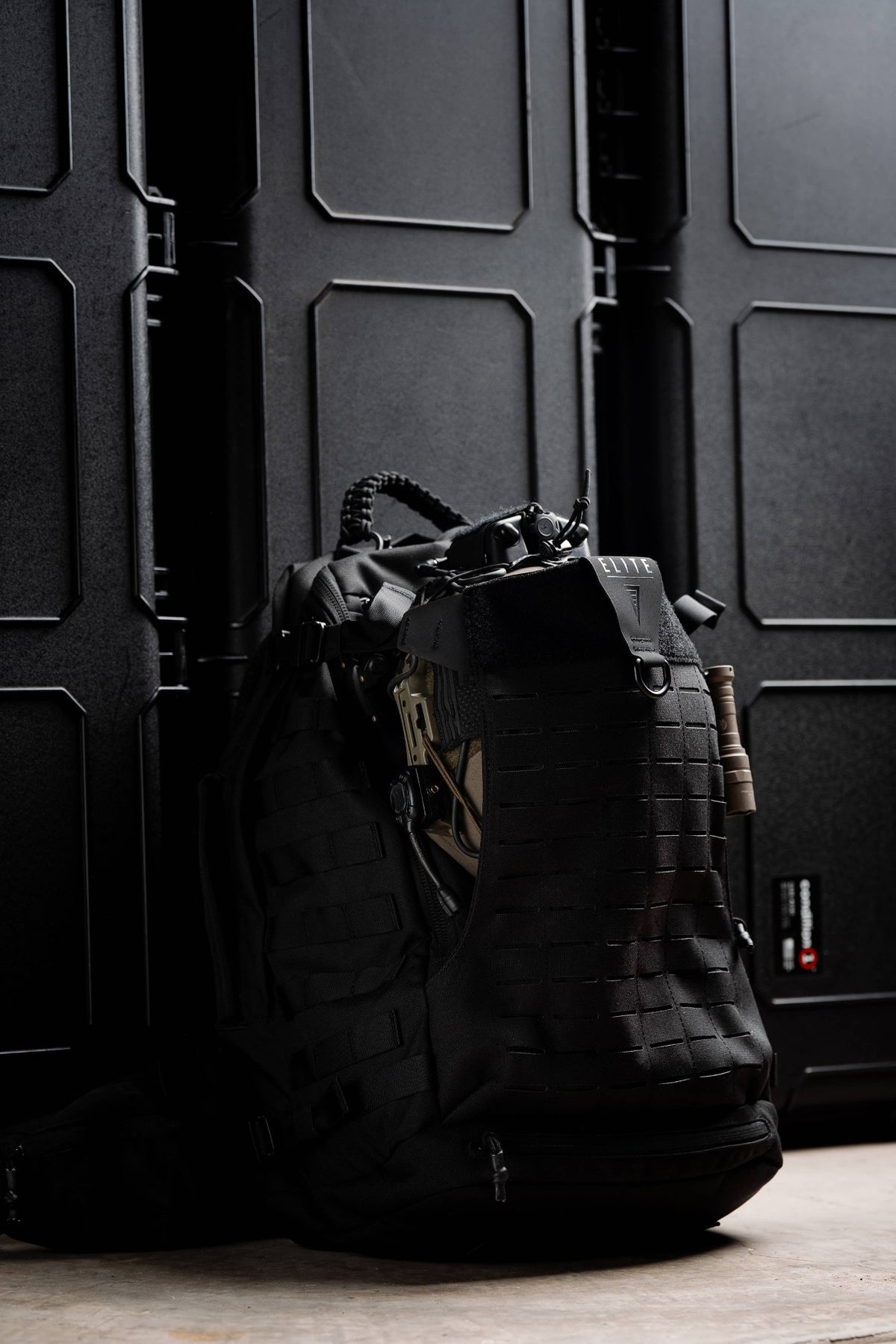 Black Mission 40L backpack with laser-cut MOLLE and ELITE logo standing in front of black hard cases