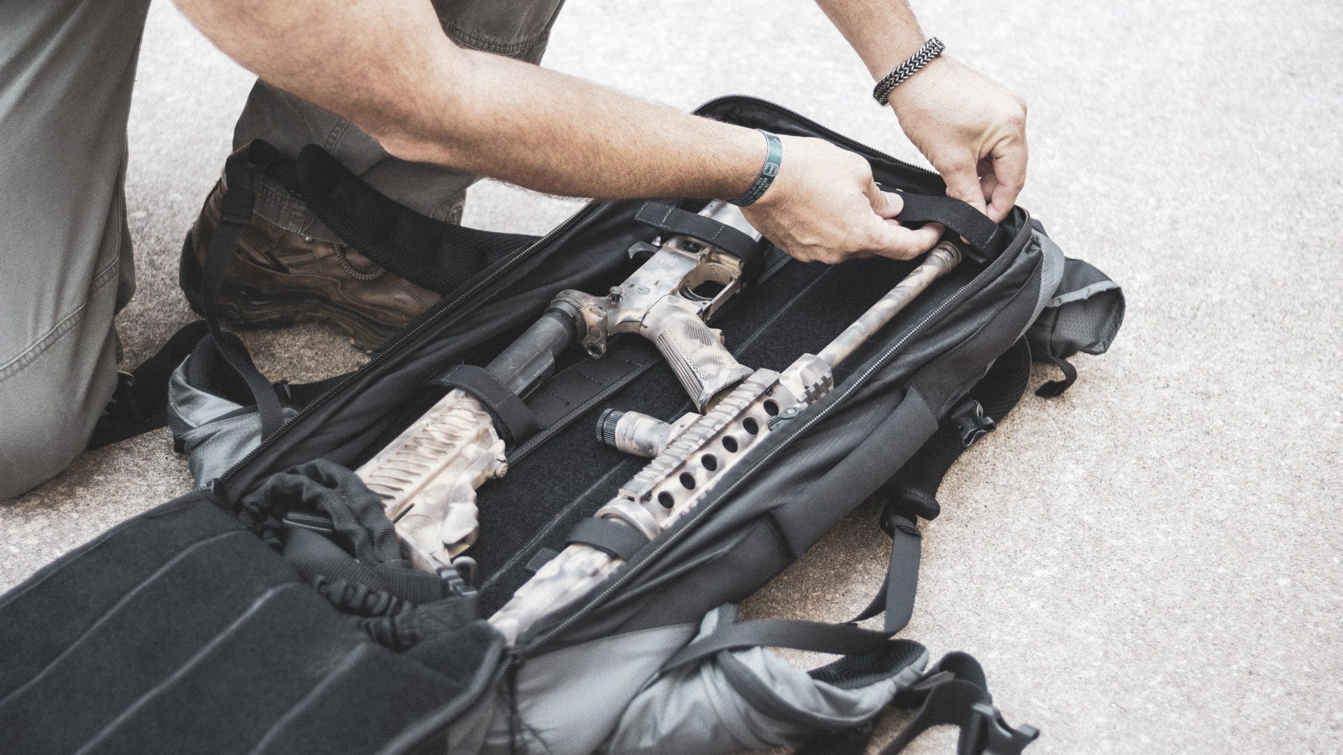 Person packing a rifle into a black backpack on a carpeted floor.